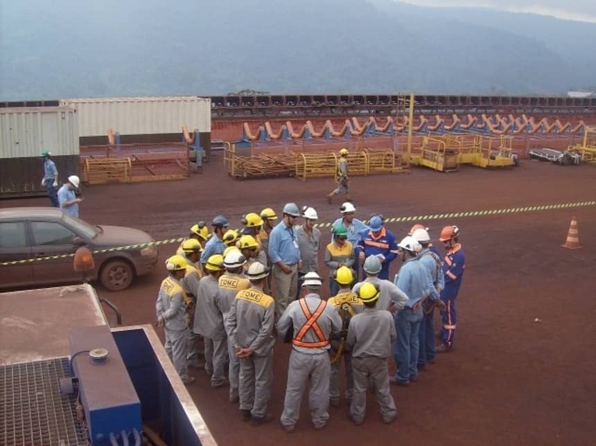 Equipe reunida em roda para treinamento de segurança ao ar livre, com instrutor ao centro em área industrial