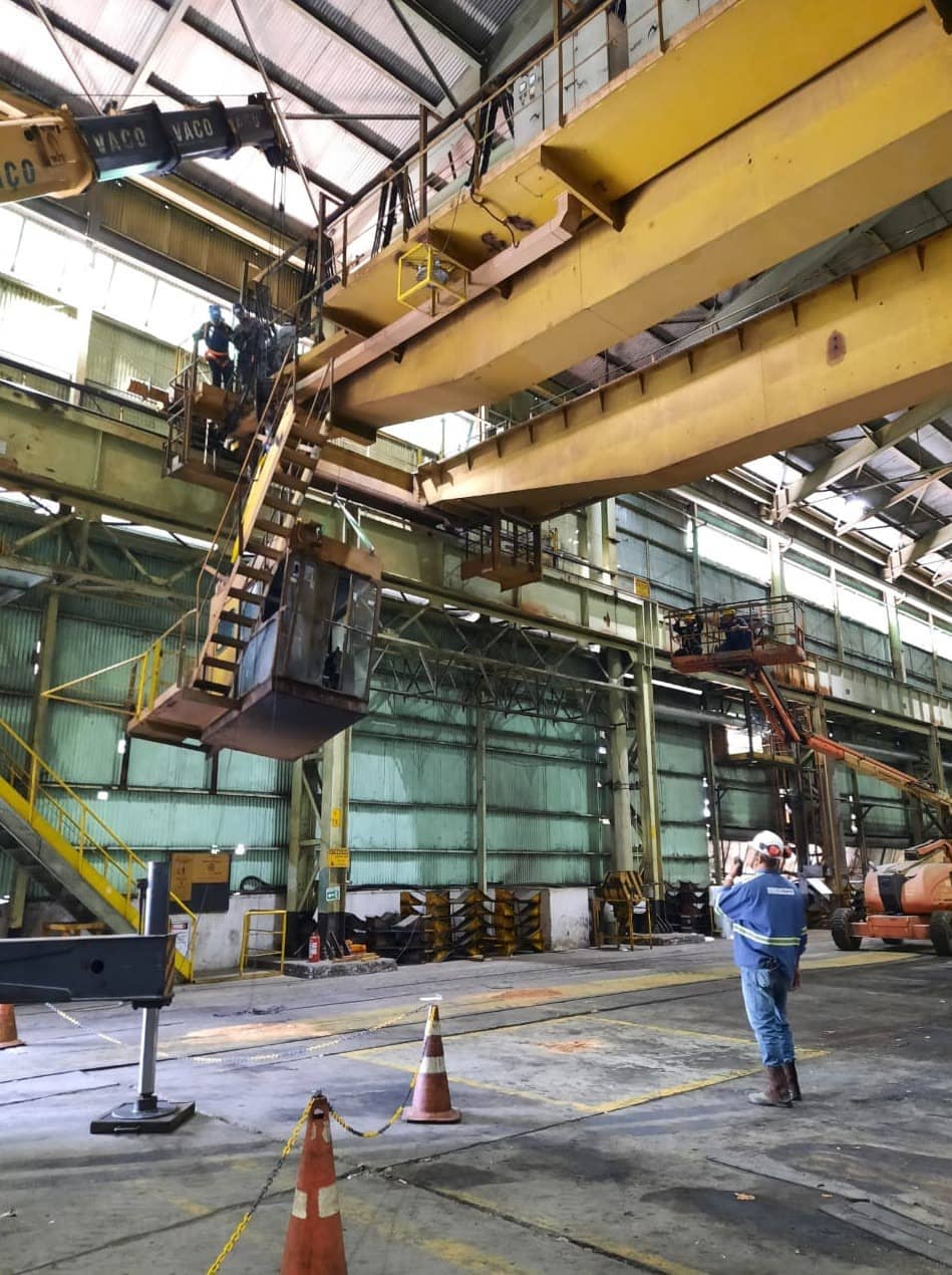 Responsável técnico com EPI observa içamento de ponte rolante em galpão industrial, com cones de sinalização e plataforma elevatória ao fundo